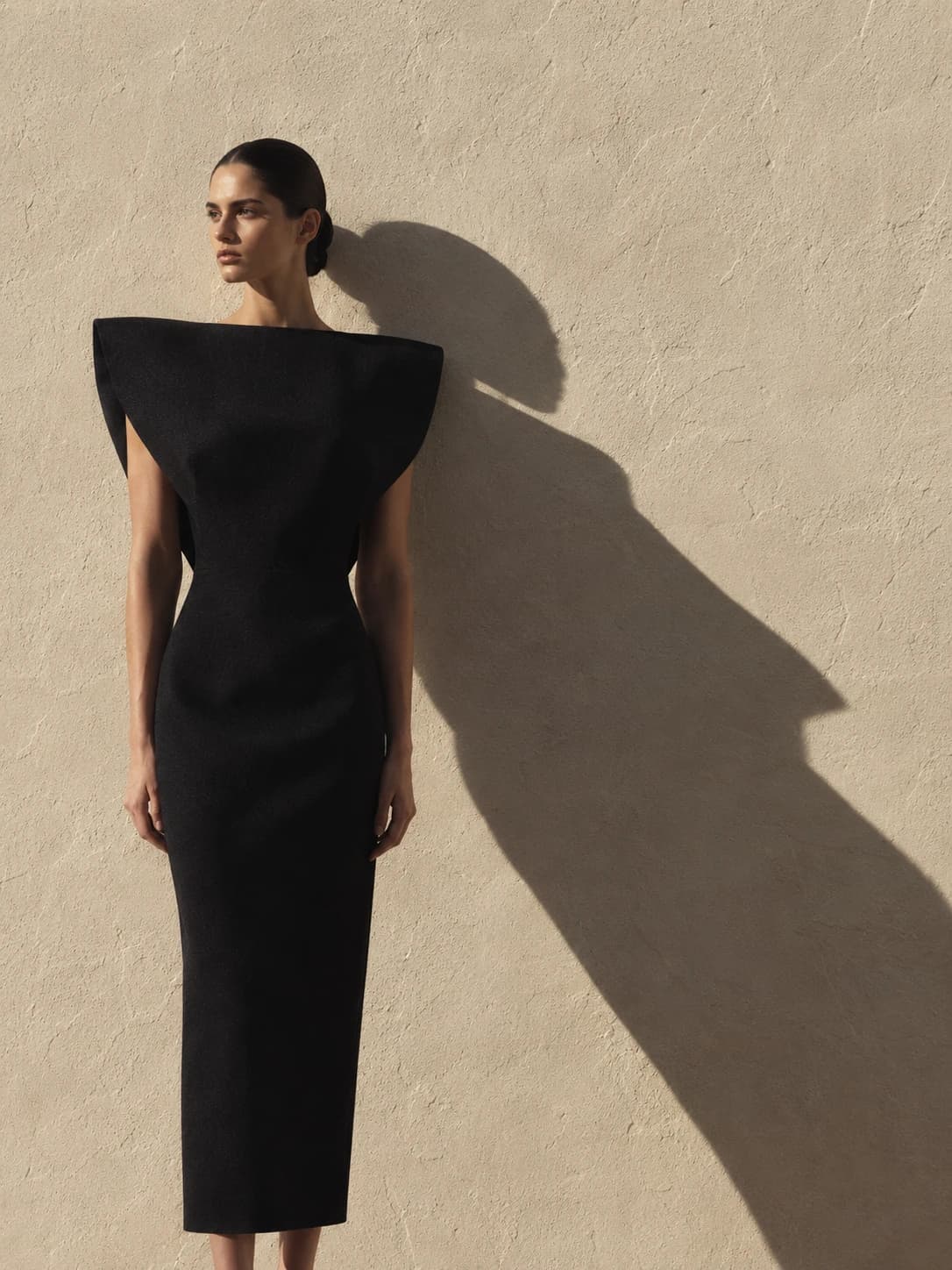 AI fashion campaign photo of a model in a sculptural black dress with exaggerated shoulders against a sunlit plaster wall, long shadow beside her.