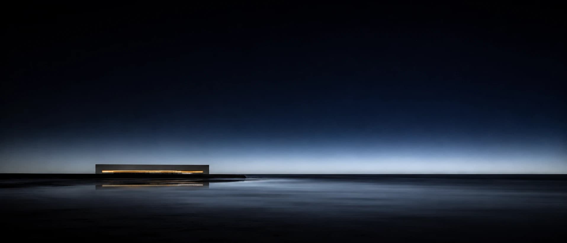 AI minimal panorama of a low concrete house with one glowing window on a flat black shoreline beneath a pale horizon at night.