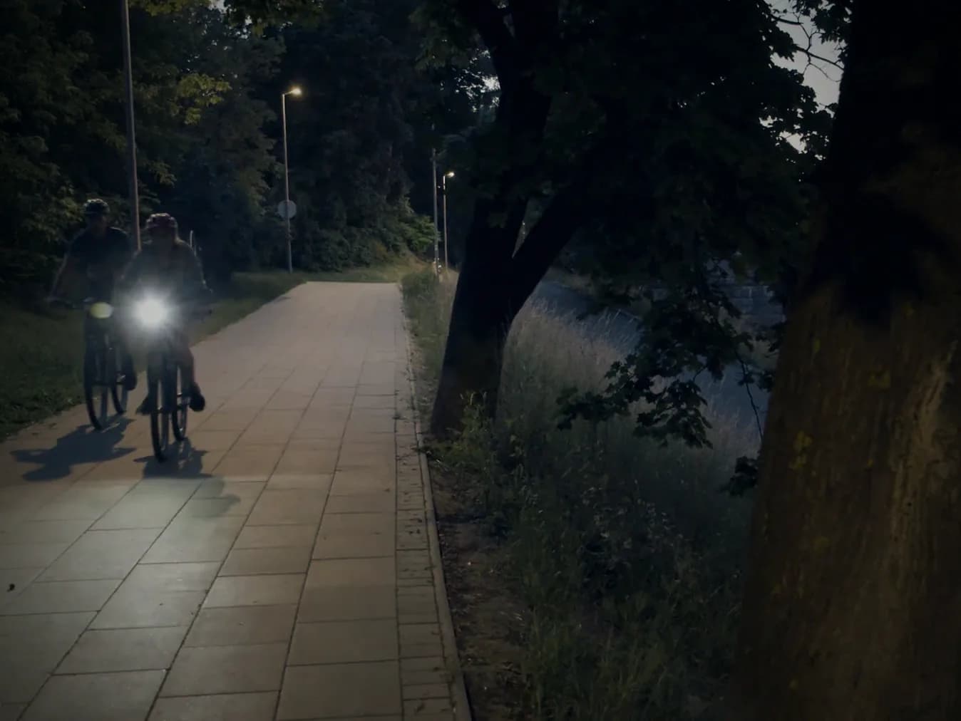 A cyclist with a bright headlamp riding along a dark tree-lined path.
