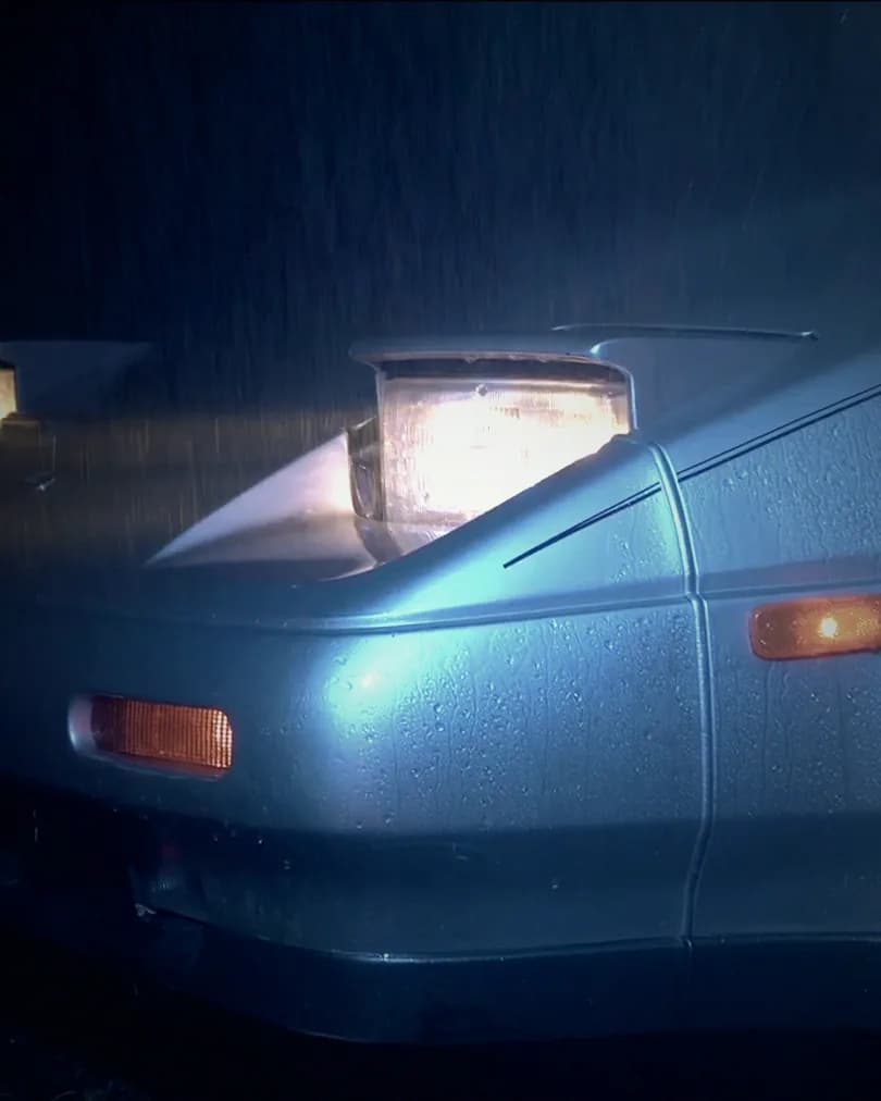 The pop-up headlight of a silver sports car glowing in the rain.